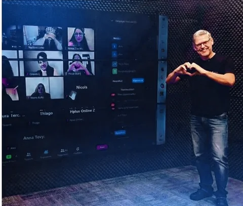 Professor fazendo um coração com as mãos ao lado da televisão com a turma na chamada