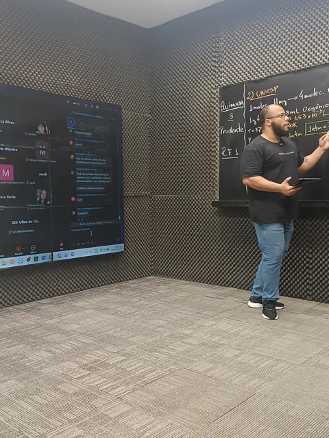Televisão do estúdio com a turma na chamada e o chat aberto, ao lado do professor